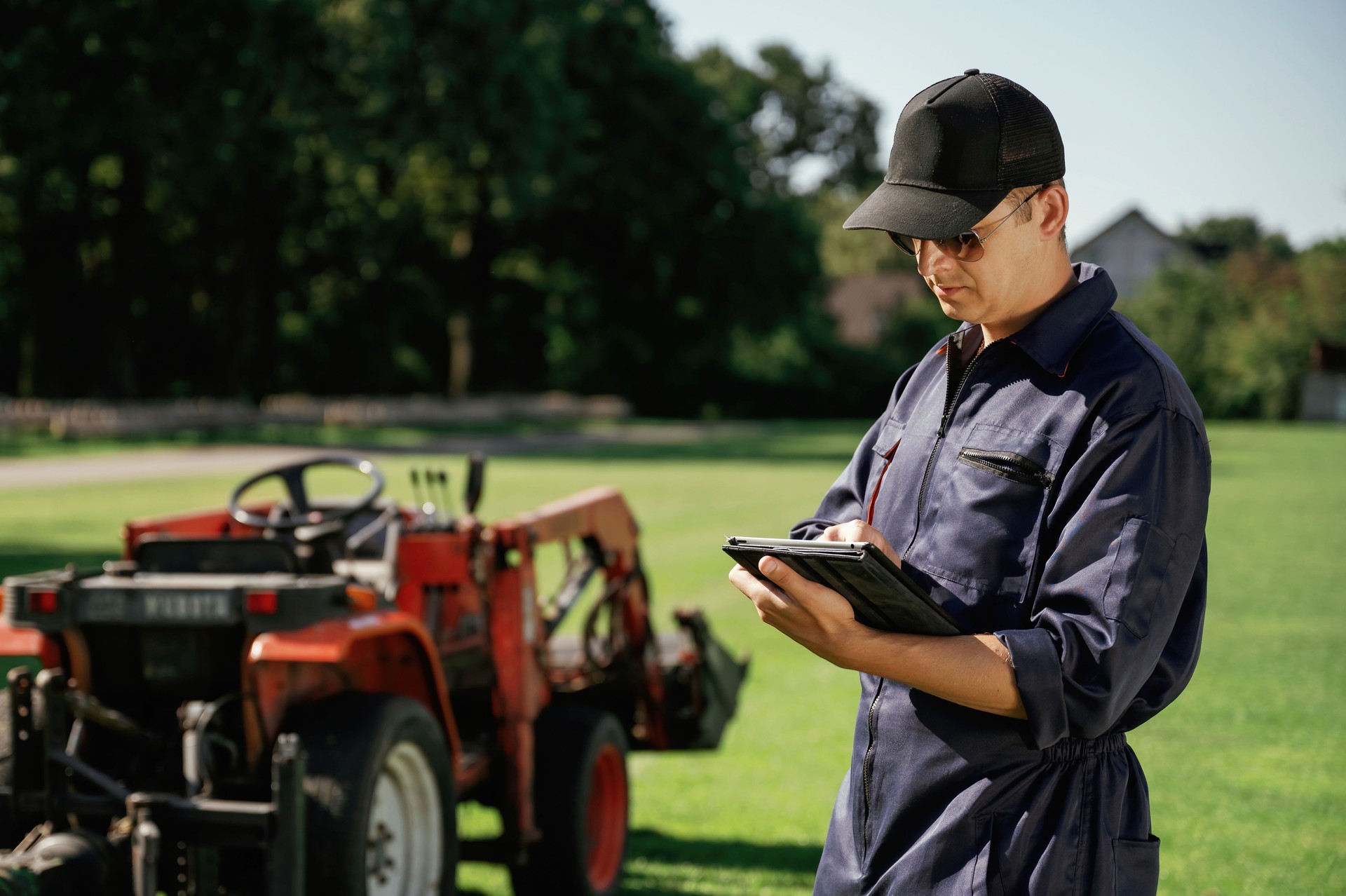 Black digital tablet in hands. Man is with utility tractor with grass cutter and aerator equipment on the field