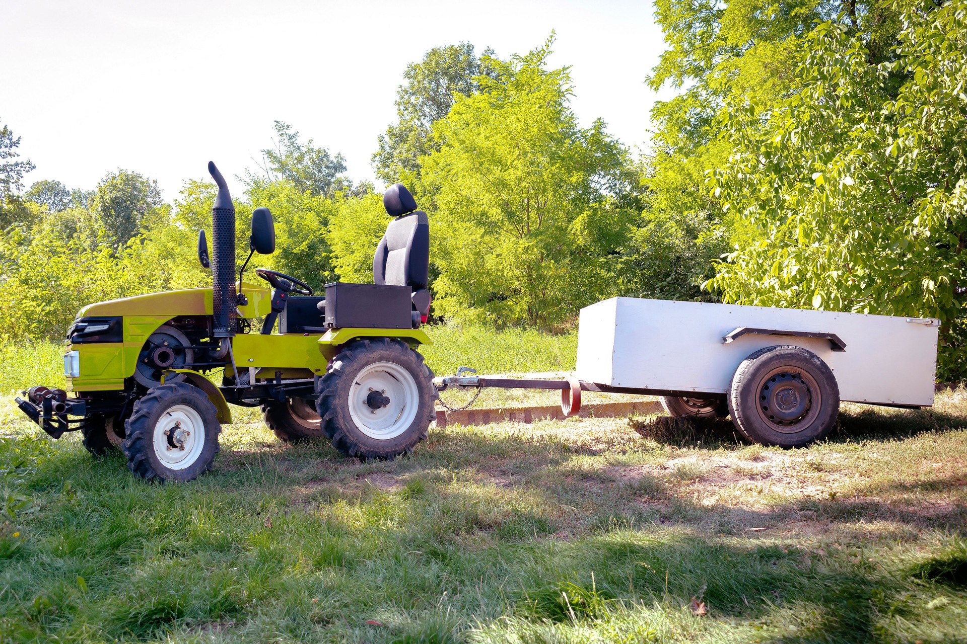Mini yellow tractor with trailer standing on field near hangar building at farm during sunny day at village area. Small agricultural machinery technology. Rural country scene